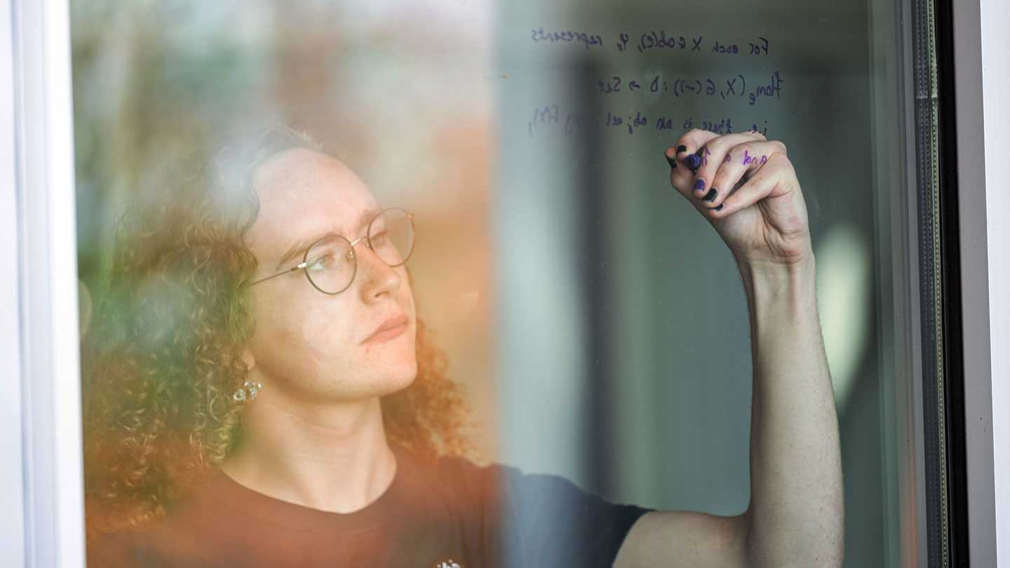 A UAH Student is writing mathematical notes on a glass window, with a blurred outdoor background.