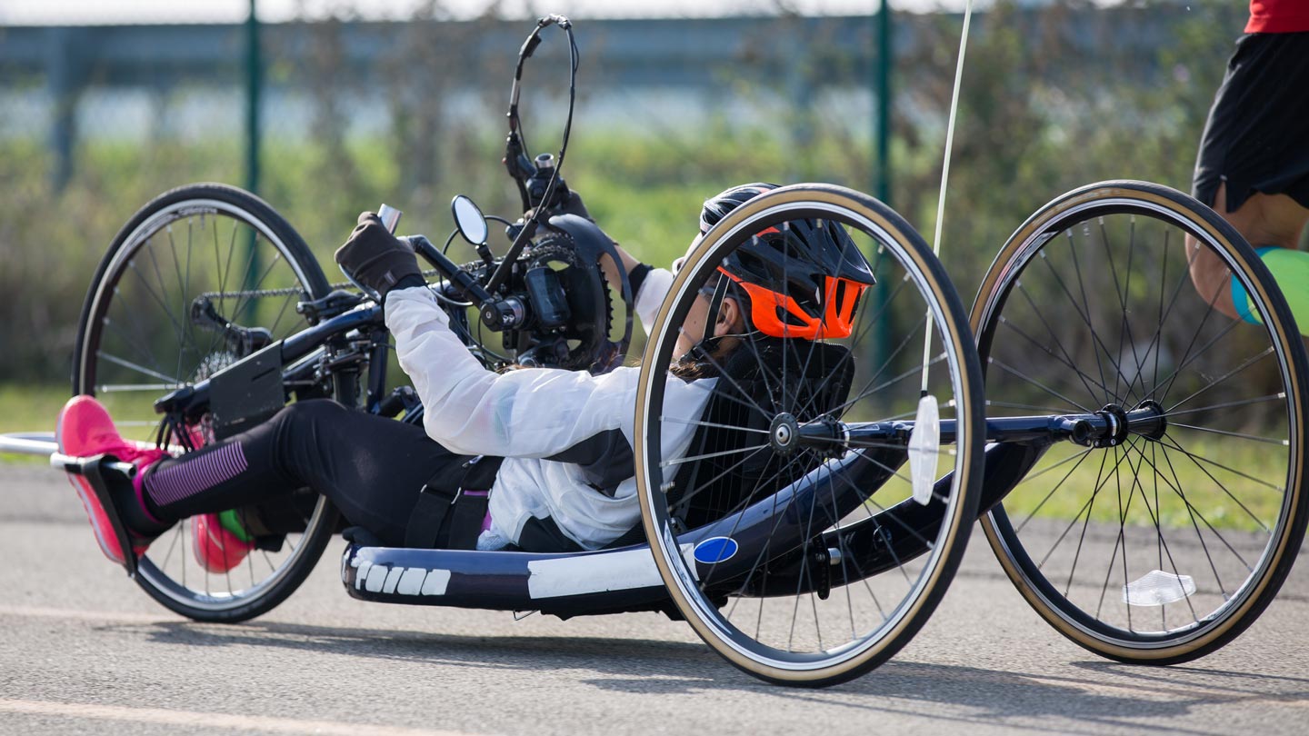Person in orange helmet riding a three-wheeled handcycle on a paved road.