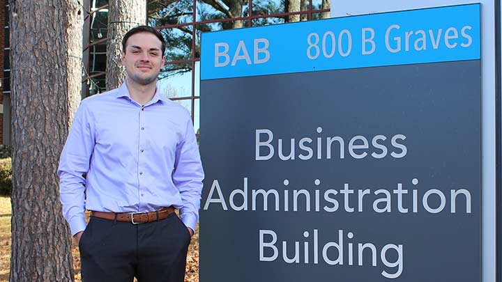 Cameron Turner outside standing by College of Business Building sign. ?>