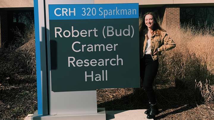 Emily Wisinski standing next to the Robert Cramer Research Hall building sign ?>