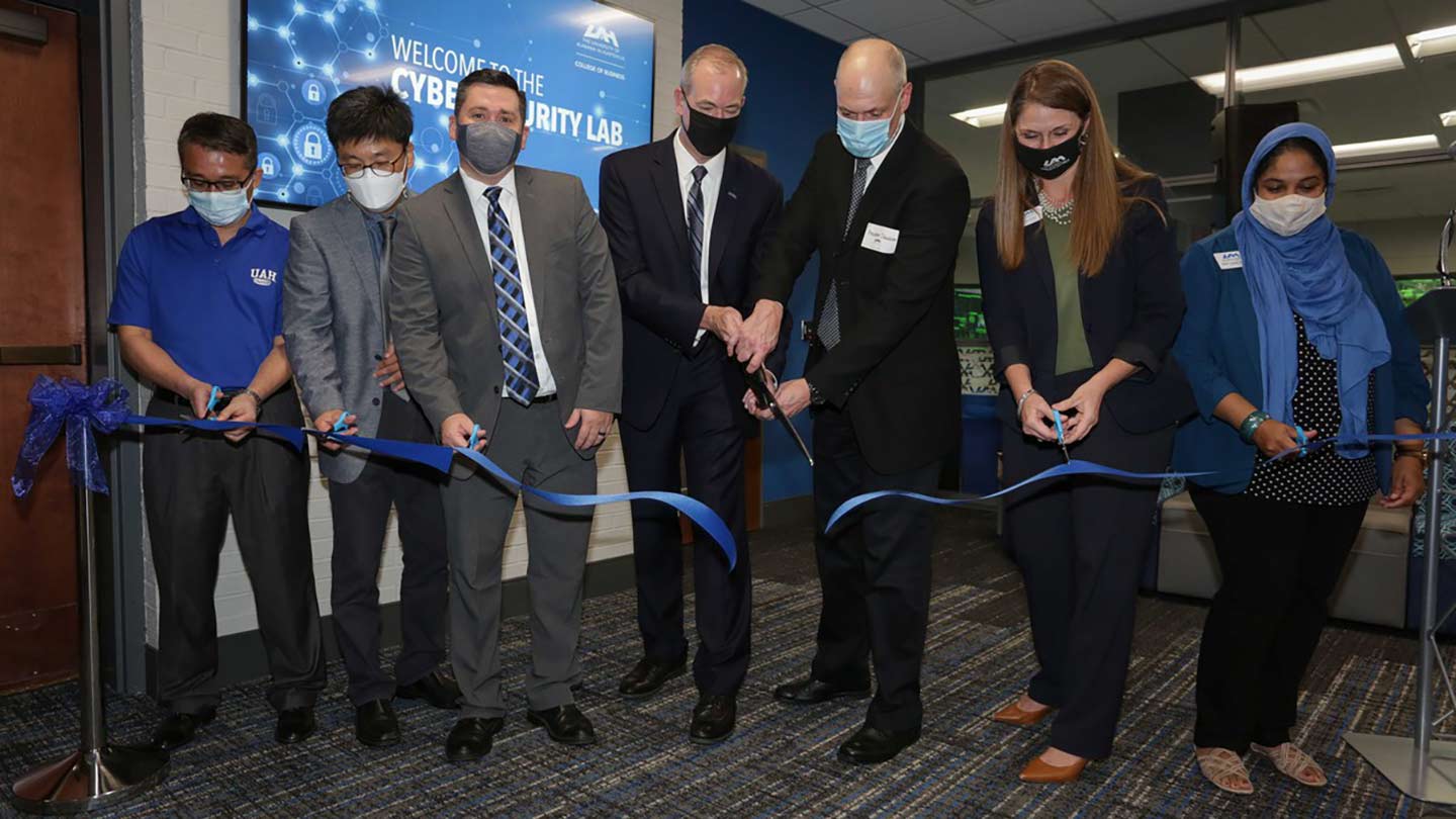 7 UAH faculty and staff members cutting a blue ribbon in front of the entrance to the new cybersecurity lab.