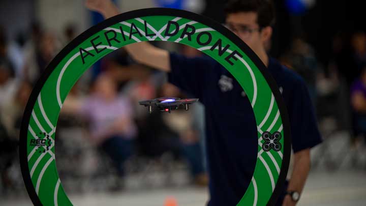 A small drone flying through a green circular gate labeled AERIAL DRONE during an indoor event with blurred audience in the background.