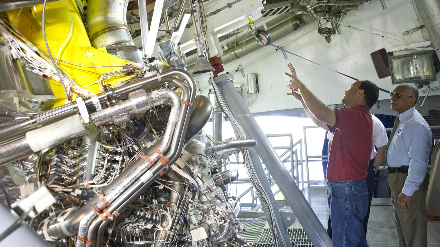 Two men observing and discussing a complex industrial machine with pipes and wires in a technical facility.
