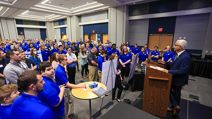 dozens of students in blue t-shirts gather around a podium where Dr. Shankar Mahalingam gives a speech