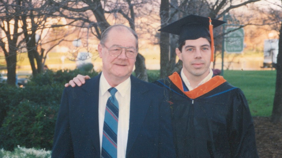 Green in cap and gown with his father, proudly smiling outdoors