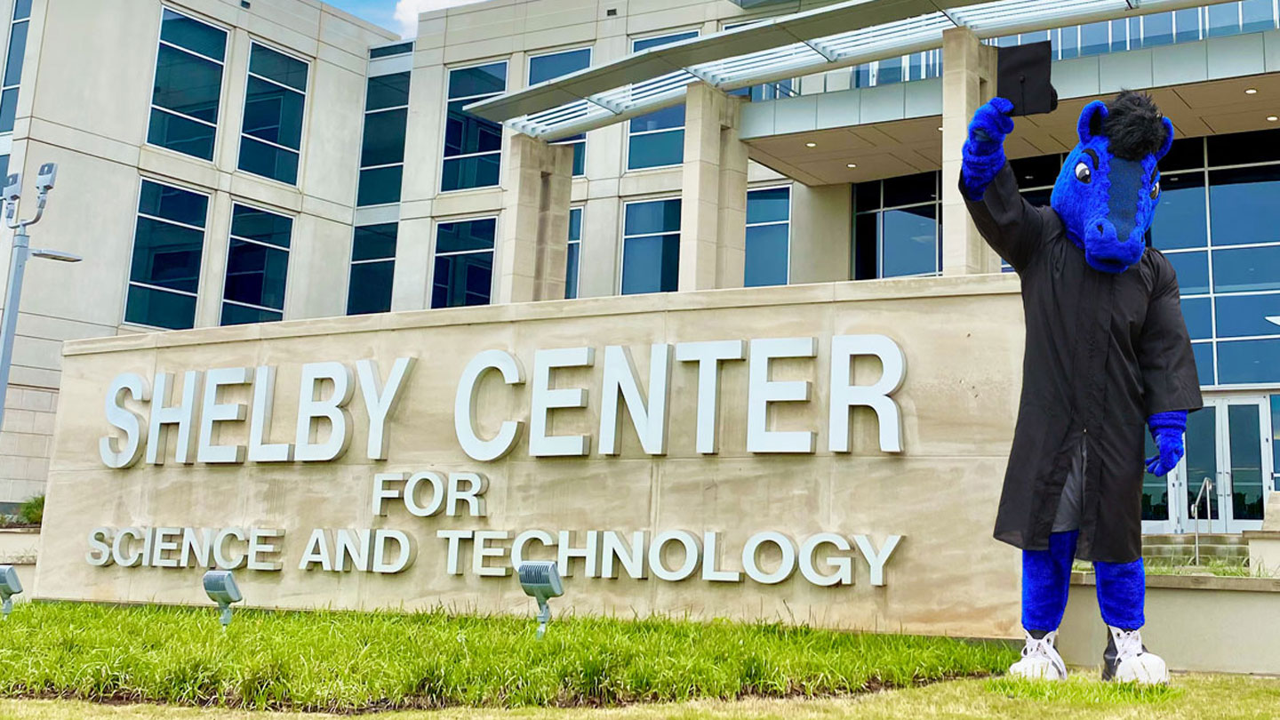 The U A H horse mascot celebrates graduation dressed in a cap and gown outside the Shelby Center for Science and Technology.