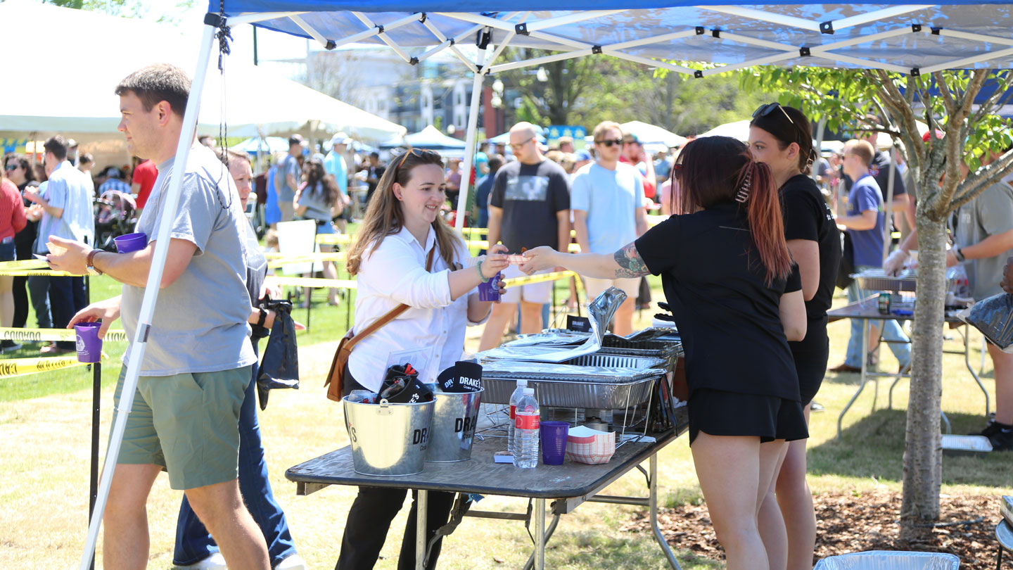 A vibrant outdoor event with people enjoying food and drinks at a service booth under a blue canopy, surrounded by a lively crowd.