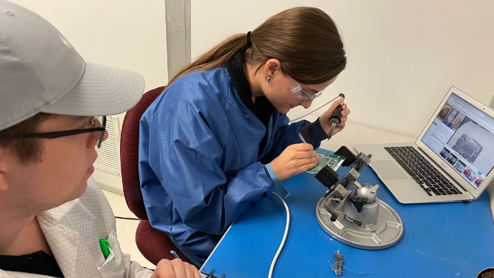 girl soldering while looking through a microscope in a lab setting with a laptop and a young man looking on