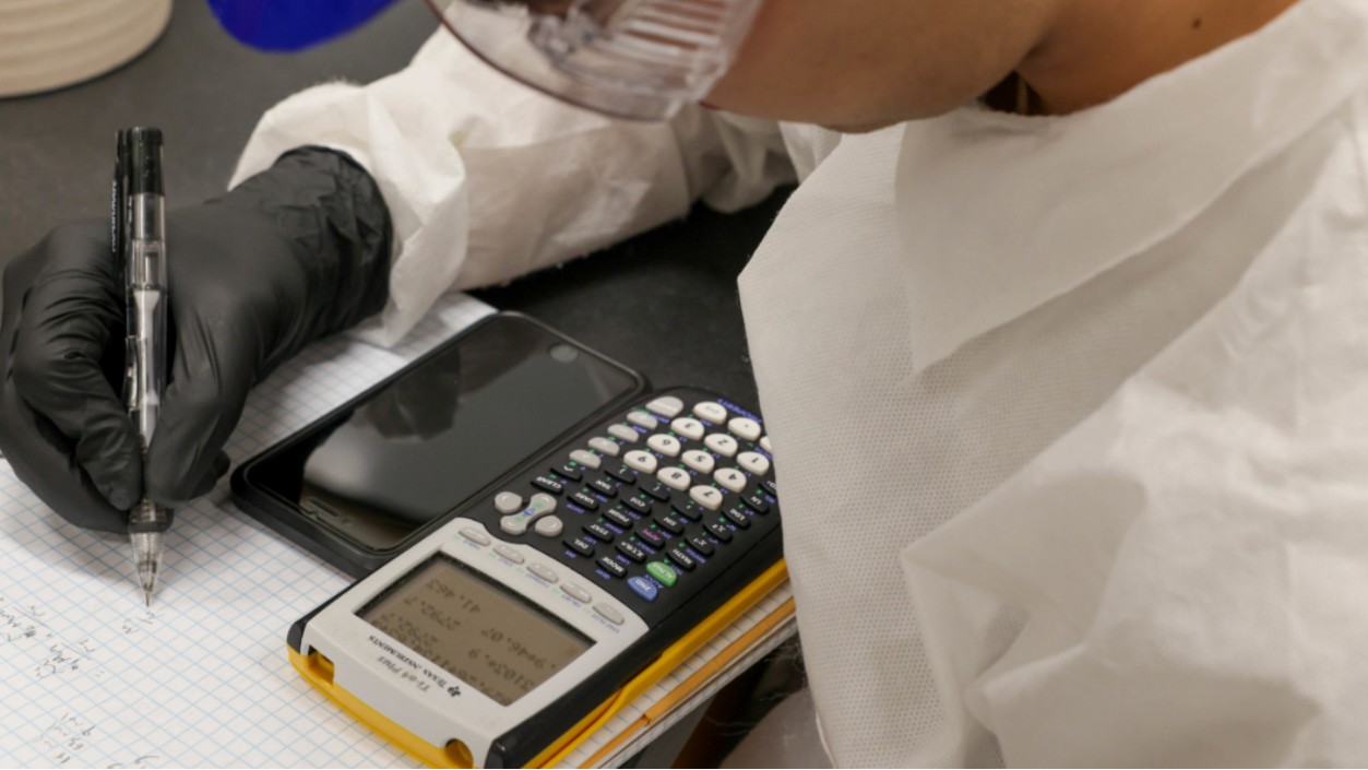 Student in lab coat sitting at a table writing on graph paper with a pencil, while using a calculator