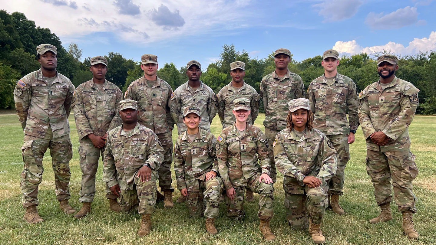  A group of eleven military personnel in uniform pose for a photo outdoors on a grassy field with trees and a partly cloudy sky in the background.