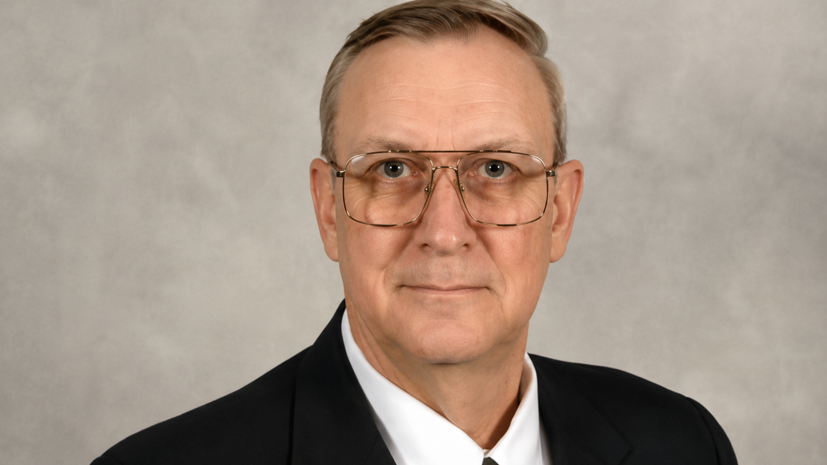 Professional headshot of Dr. William “Buddy” Bishop wearing a dark suit, white shirt, and glasses against a light gray studio background.